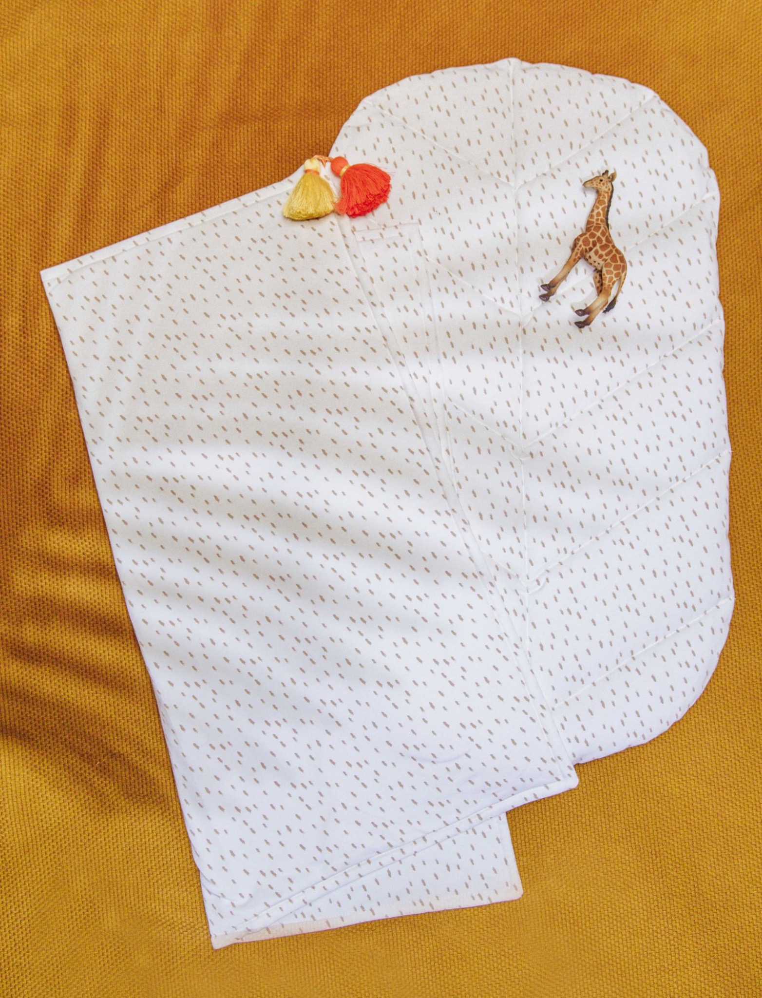 Biała kołderka dziecięca w brązowe kropki, zdobiona żyrafą i pomponami, leży na musztardowym tle. Delikatne światło podkreśla detale wzoru.