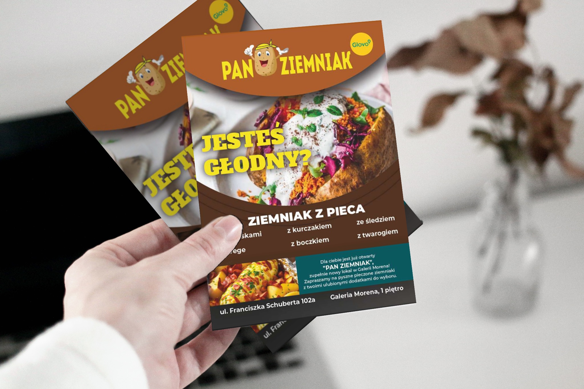 Dwa ulotki reklamowe restauracji 'Pan Ziemniak' prezentowane w dłoni, z grafiką pieczonego ziemniaka z dodatkami i informacją o lokalizacji w Galerii Morena.
