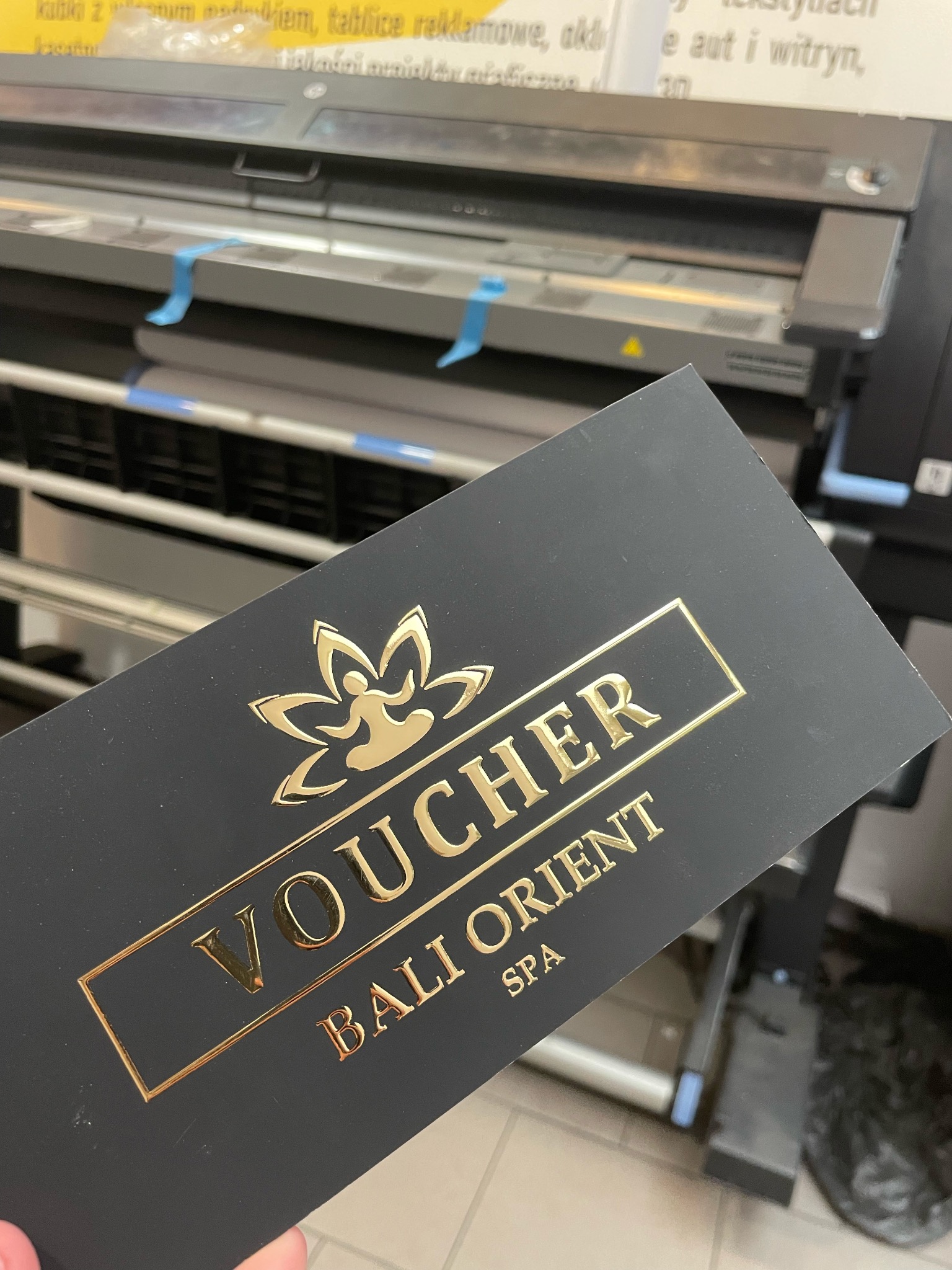 Czarny voucher Bali Orient Spa z tłoczonym złotym napisem, leżący na tle wielkoformatowej drukarki