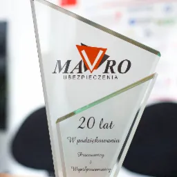 Szklana statuetka z logo firmy ubezpieczeniowej Mavro, upamiętniająca 20-lecie działalności, z dedykacją dla pracowników i współpracowników z roku 2019, na drewnianym biurku.