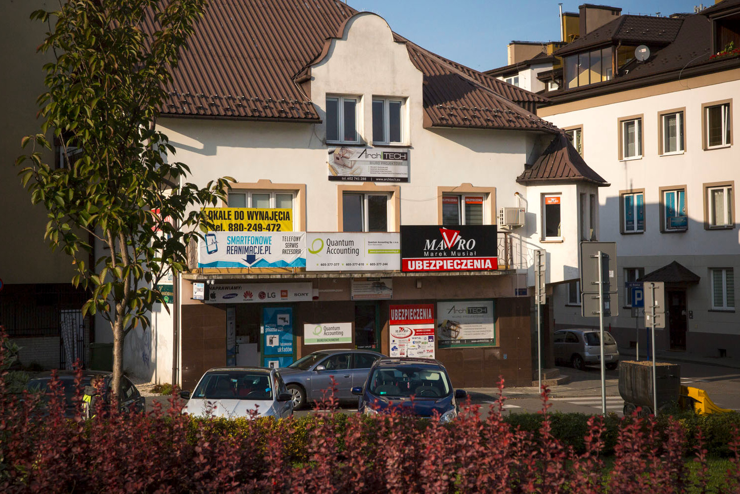 Budynek usługowy z szyldami różnych firm, w tym z logo firmy ubezpieczeniowej 'Mavro' z napisem 'Ubezpieczenia', widok z poziomu ulicy przez krzewy.