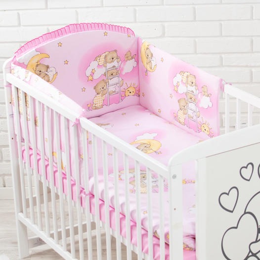 Łóżeczko dziecięce z białymi szczebelkami i różową pościelą w misie, księżyce i gwiazdki na tle białej ceglanej ściany.