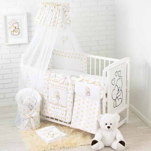 Białe łóżeczko dziecięce z baldachimem i pościelą w złote gwiazdki, pluszowy miś i obrazek z motywem misia na tle białej cegły.