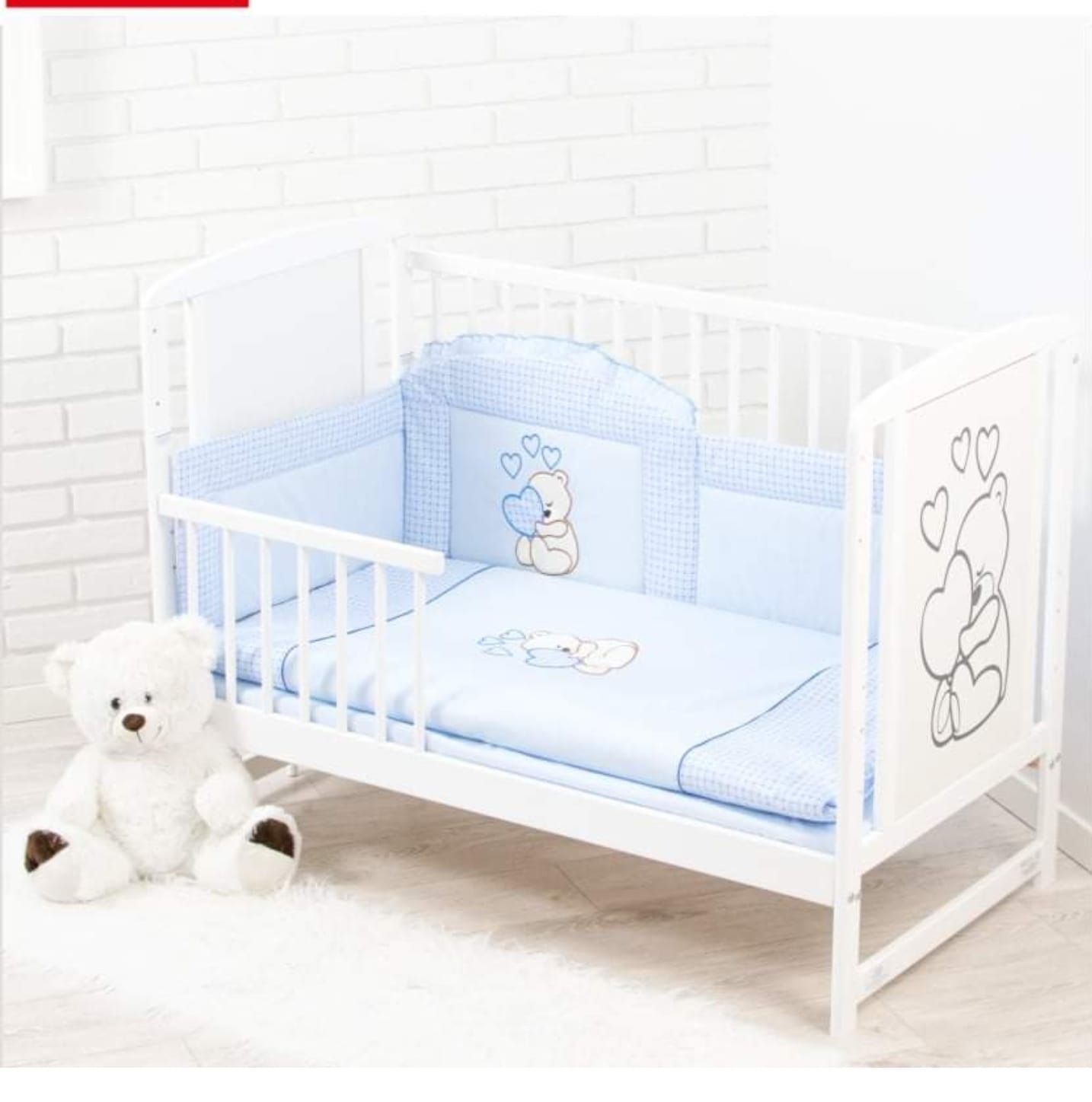 Białe łóżeczko niemowlęce z niebieską pościelą w kratkę i motywem misia, pluszowy miś siedzący obok na białym dywanie.