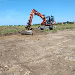 Pomarańczowa koparka Atlas na kołach, z łyżką, przygotowuje teren pod budowę, widoczne ślady opon na ziemi i trawa w tle.