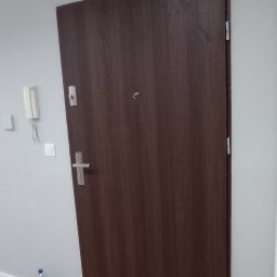 JACO Bud&Art Jacek Kozina - Ciemne, drewnopodobne drzwi wejściowe w szarym korytarzu z panelami na podłodze i białą skrzynką elektryczną na ścianie.