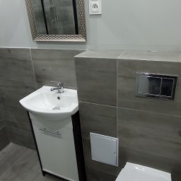 JACO Bud&Art Jacek Kozina - Nowoczesna łazienka z szarymi płytkami imitującymi drewno, białą umywalką z szafką, toaletą i lustrem w ozdobnej ramie, w którym odbija się kabina prysznicowa.