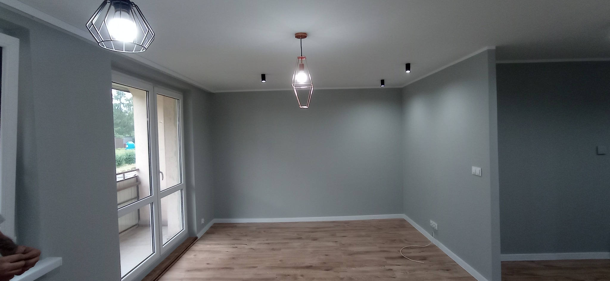 Wnętrze pokoju po remoncie: szare ściany, panele podłogowe, okno balkonowe i designerskie lampy wiszące