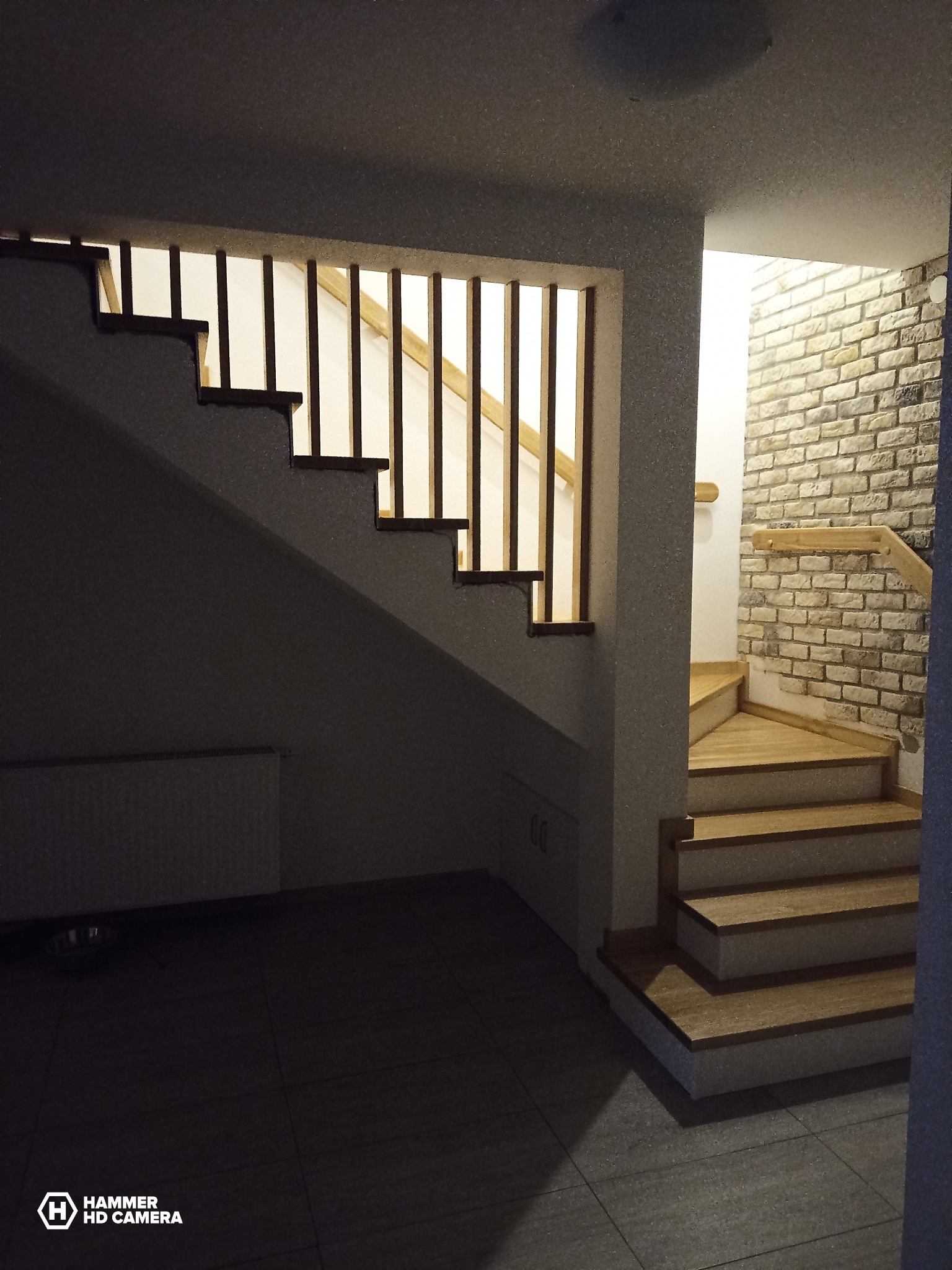 Fragment klatki schodowej z drewnianymi schodami i ażurową balustradą, widoczne schody prowadzące w górę z drewnianymi stopniami i ceglaną ścianą w tle.