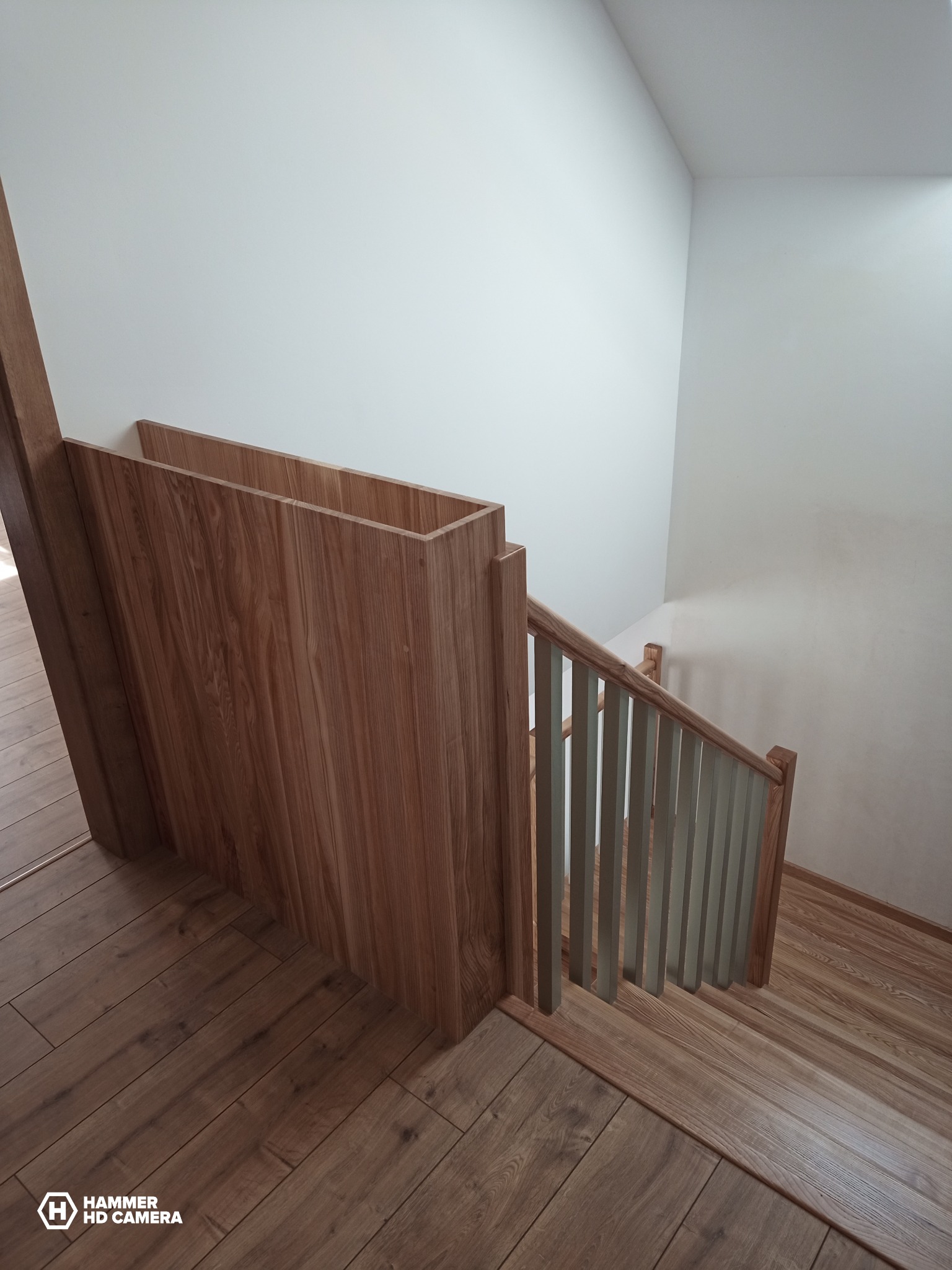 Wnętrze domu z widokiem na drewniane schody z jasnymi, pionowymi szczeblami balustrady, drewnianą podłogą i białymi ścianami.