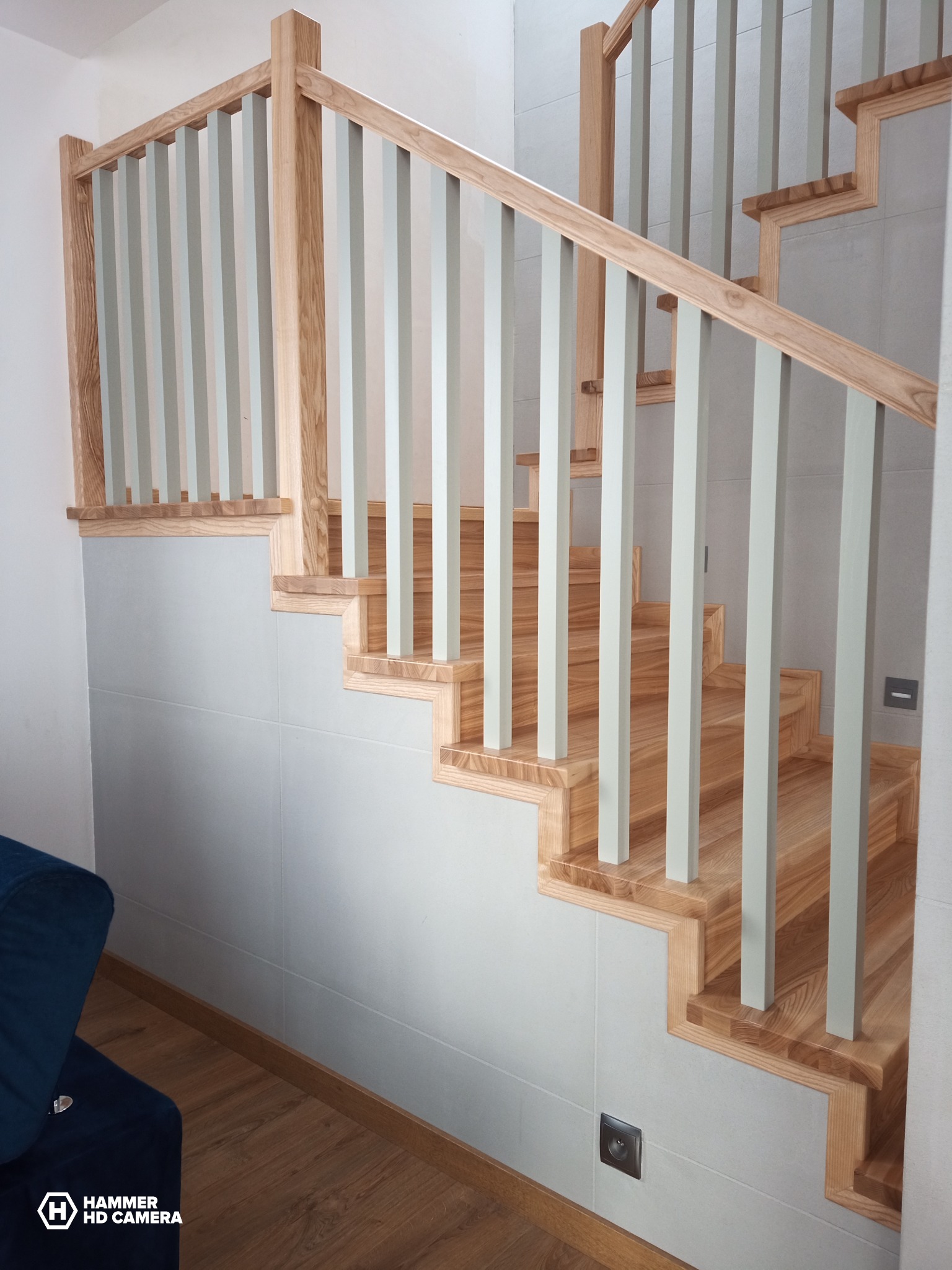 Jasne, drewniane schody z pastelowymi, szarymi balustradami w nowoczesnym wnętrzu, widok z dołu na pierwsze stopnie i fragment niebieskiej kanapy.