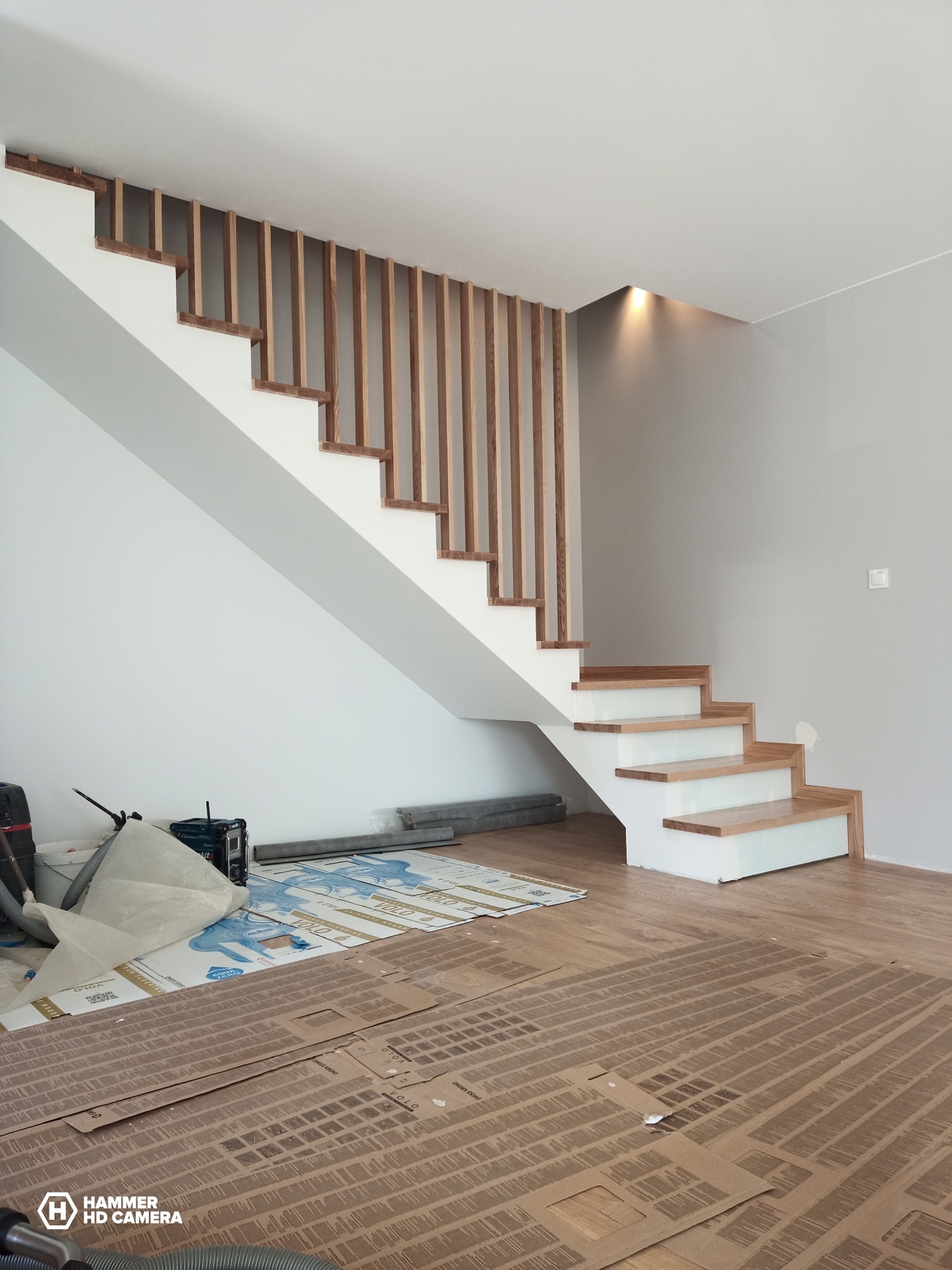 Nowoczesne schody z drewnianymi stopniami i pionowymi balustradami, białe ściany, ochrona podłogi kartonem podczas montażu.