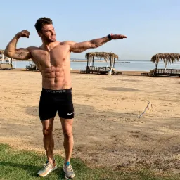 Mężczyzna w sportowych spodenkach Nike Pro prezentuje swoją muskulaturę na piaszczystej plaży z widokiem na morze i palapami.