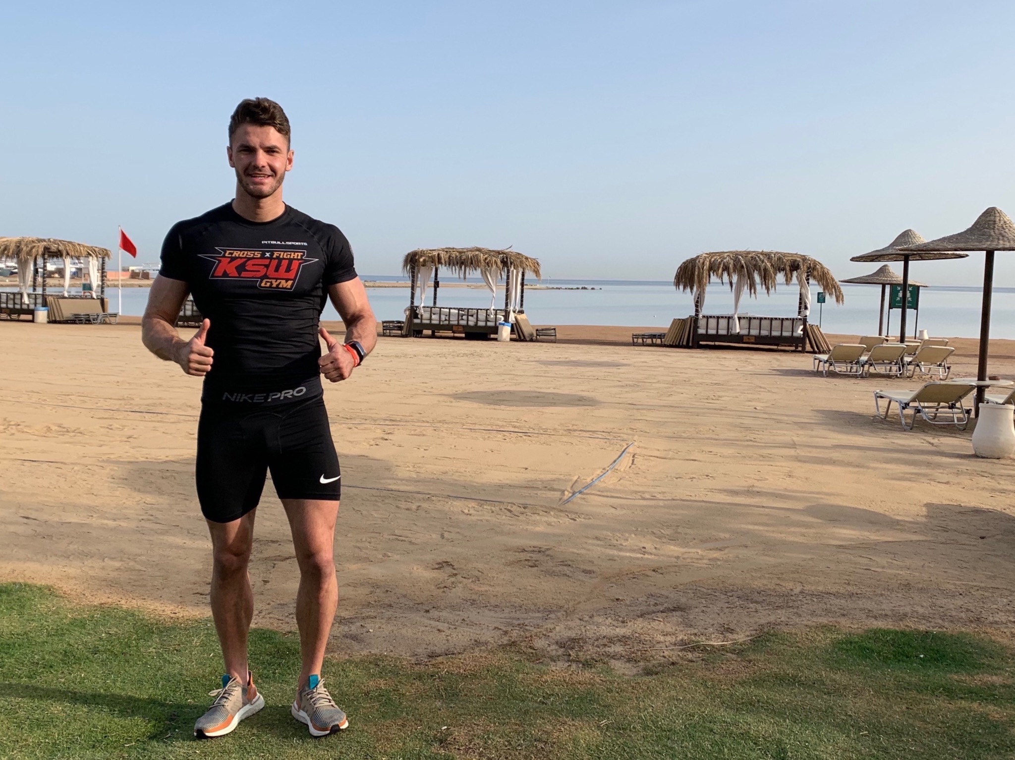 Uśmiechnięty mężczyzna w sportowym stroju na plaży, z podniesionymi kciukami, w tle widoczne palmy i leżaki.