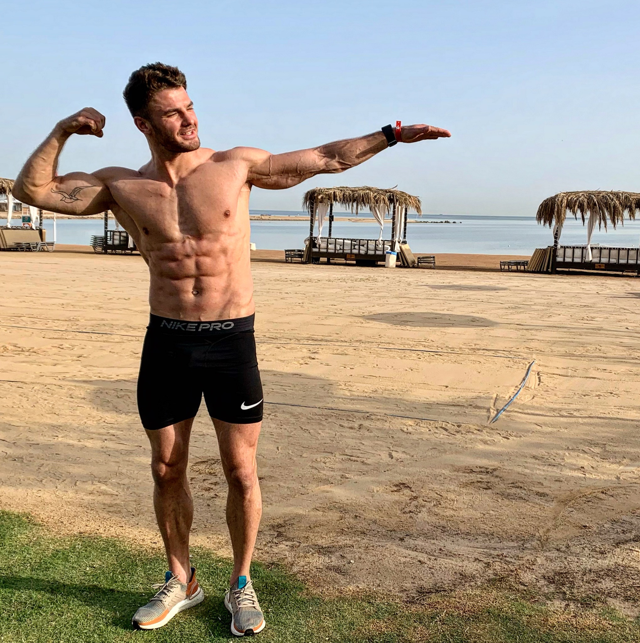 Mężczyzna w sportowych spodenkach Nike Pro prezentuje swoją muskulaturę na piaszczystej plaży z widokiem na morze i palapami.