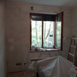 Usługi Remontowo Malarskie - Pokój w trakcie przygotowania do malowania: ściany po nałożeniu gładzi, okno z roletami otwarte na oścież, drabina oparta o ścianę, meble przykryte folią ochronną, wiadro z farbą na podłodze.