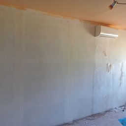 Usługi Remontowo Malarskie - Przygotowanie ściany do malowania: częściowo zagruntowana ściana, zabezpieczona folią klimatyzacja i drabina oparta o ścianę przy oknie.