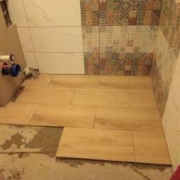 Usługi Remontowo Malarskie - Układanie drewnopodobnych płytek podłogowych w łazience, widoczne fragmenty ściany z dekoracyjnymi i białymi płytkami oraz niezakończone prace przy instalacji sanitarnej.