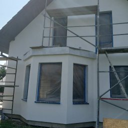 Usługi Remontowo Malarskie - Dom jednorodzinny z oknami zabezpieczonymi folią i taśmą malarską, otoczony rusztowaniem podczas prac elewacyjnych w słoneczny dzień.