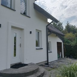Usługi Remontowo Malarskie - Biały dom z nową elewacją, widoczne okna z roletami, czarne rynny, schody wejściowe z czarnym materiałem i fragment ogrodu z roślinami.