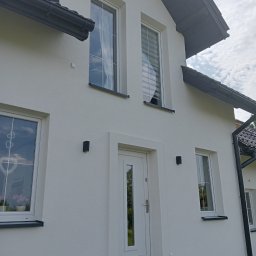 Usługi Remontowo Malarskie - Biała fasada domu jednorodzinnego z widocznymi oknami z białymi ramami, drzwiami wejściowymi i czarnymi lampami elewacyjnymi, po zakończonym remoncie.