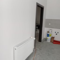 Usługi Remontowo Malarskie - Świeżo pomalowane na biało ściany w pokoju, widoczny biały grzejnik, szara podłoga, czarna ościeżnica drzwi i wiaderka z farbą w tle.