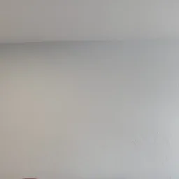 Ściana wewnętrzna pomalowana na jasny odcień szarości z widoczną fakturą, obok framuga okienna z niebieską taśmą malarską.