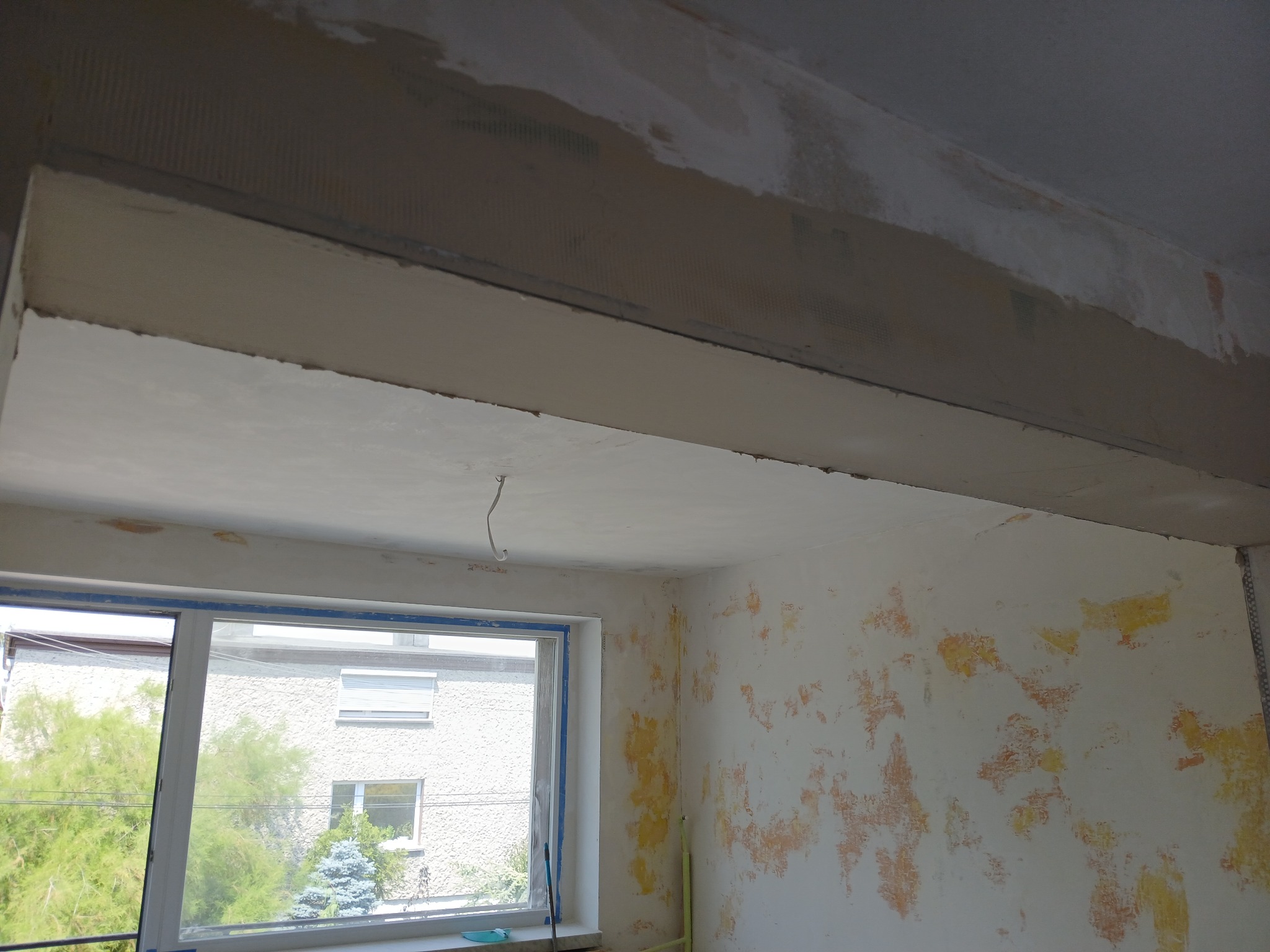 Wnętrze pokoju w trakcie remontu: ściany z resztkami starej farby, okno zabezpieczone taśmą malarską, sufit z nałożoną warstwą gładzi, widoczny kabel elektryczny.