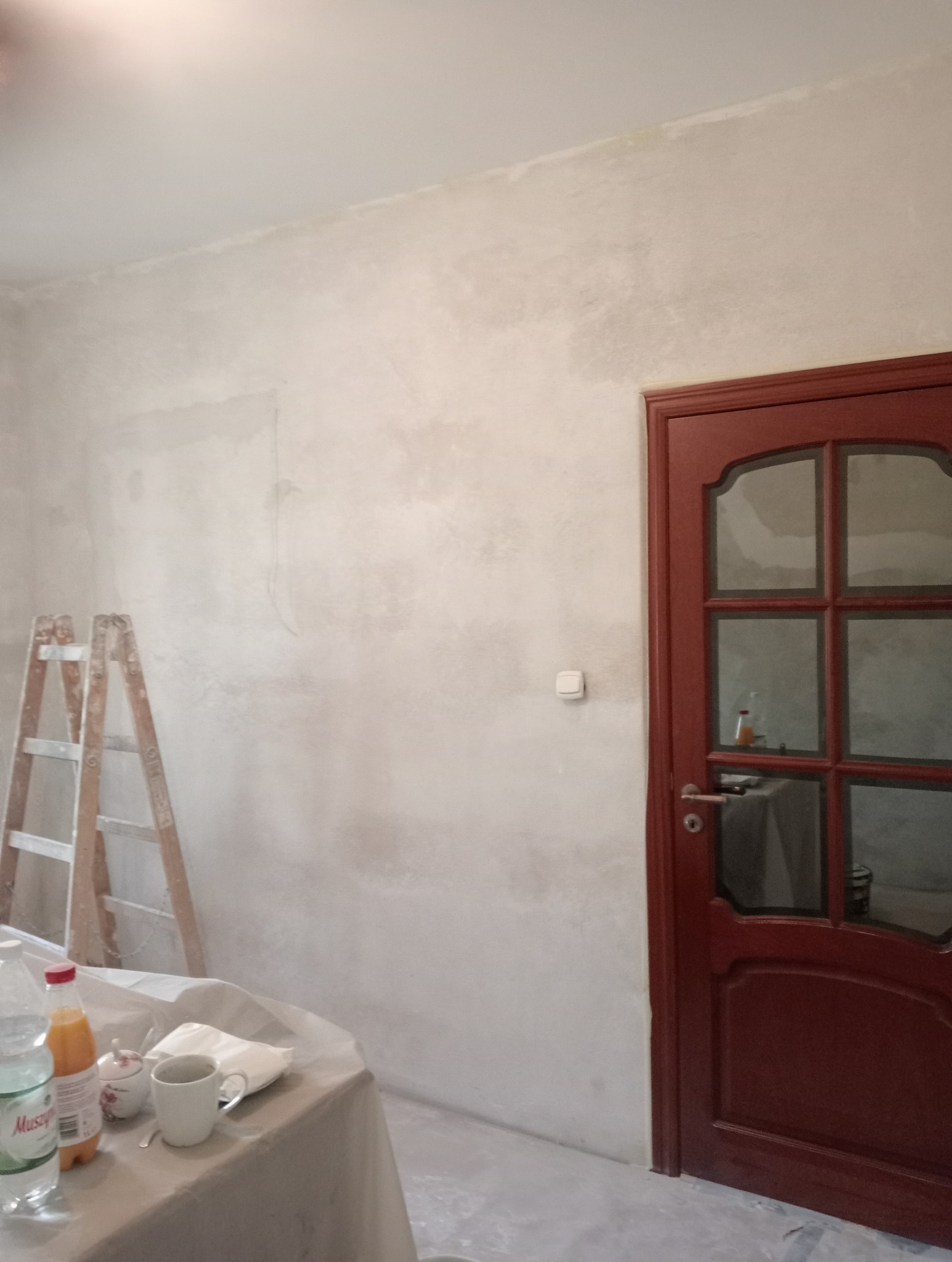 Ściana po nałożeniu gładzi, przed szlifowaniem, z widoczną drabiną, przykrytym stołem z napojami i ciemnymi drzwiami z szybami