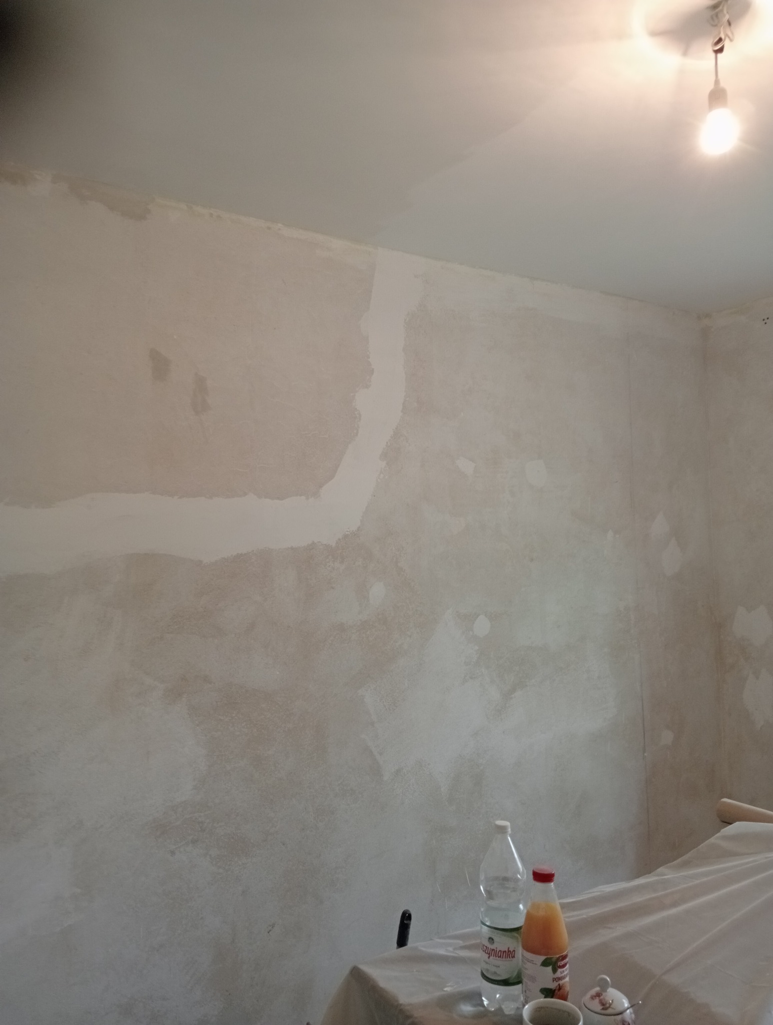 Ściana w trakcie szpachlowania, widoczne nierówności i białe fragmenty szpachli, zabezpieczone meble pod ścianą.