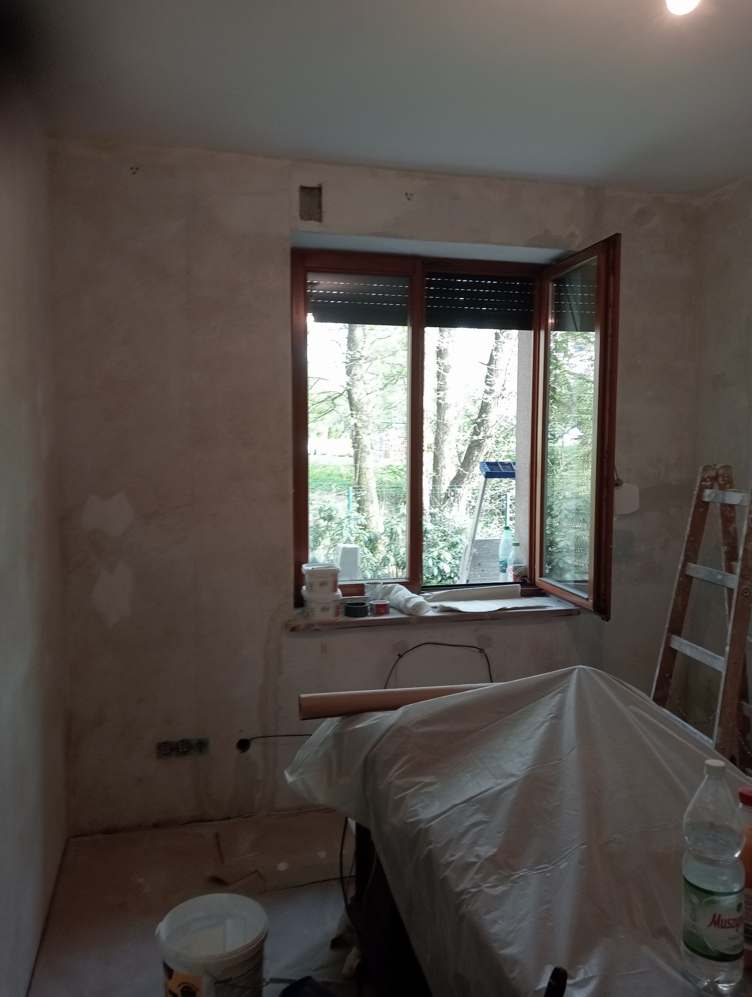 Pokój w trakcie przygotowania do malowania: ściany po nałożeniu gładzi, okno z roletami otwarte na oścież, drabina oparta o ścianę, meble przykryte folią ochronną, wiadro z farbą na podłodze.