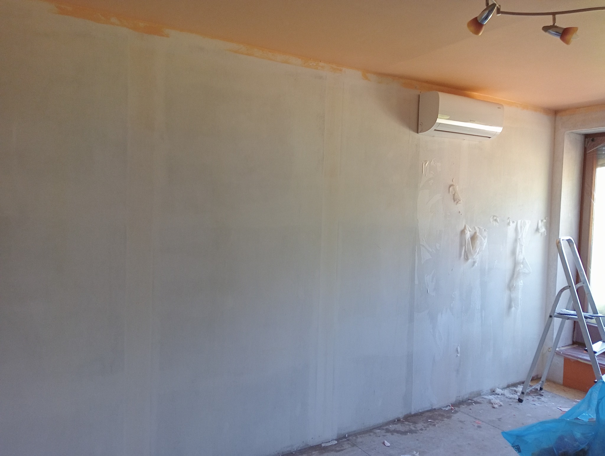 Przygotowanie ściany do malowania: częściowo zagruntowana ściana, zabezpieczona folią klimatyzacja i drabina oparta o ścianę przy oknie.