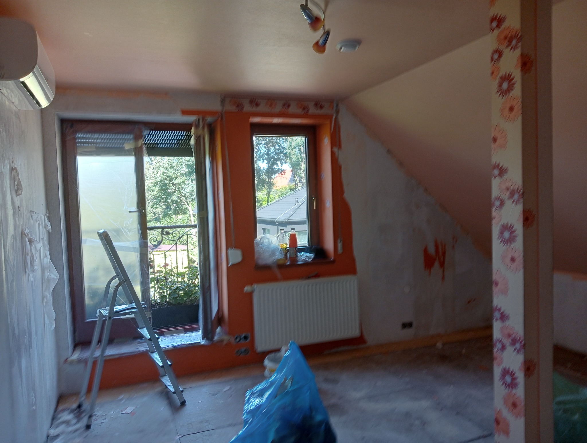 Pokój na poddaszu w trakcie remontu: ściany z resztkami starej farby, okna zabezpieczone folią, drabina oparta o okno balkonowe, grzejnik, butelki na parapecie, kolumna z kwiatowym wzorem...