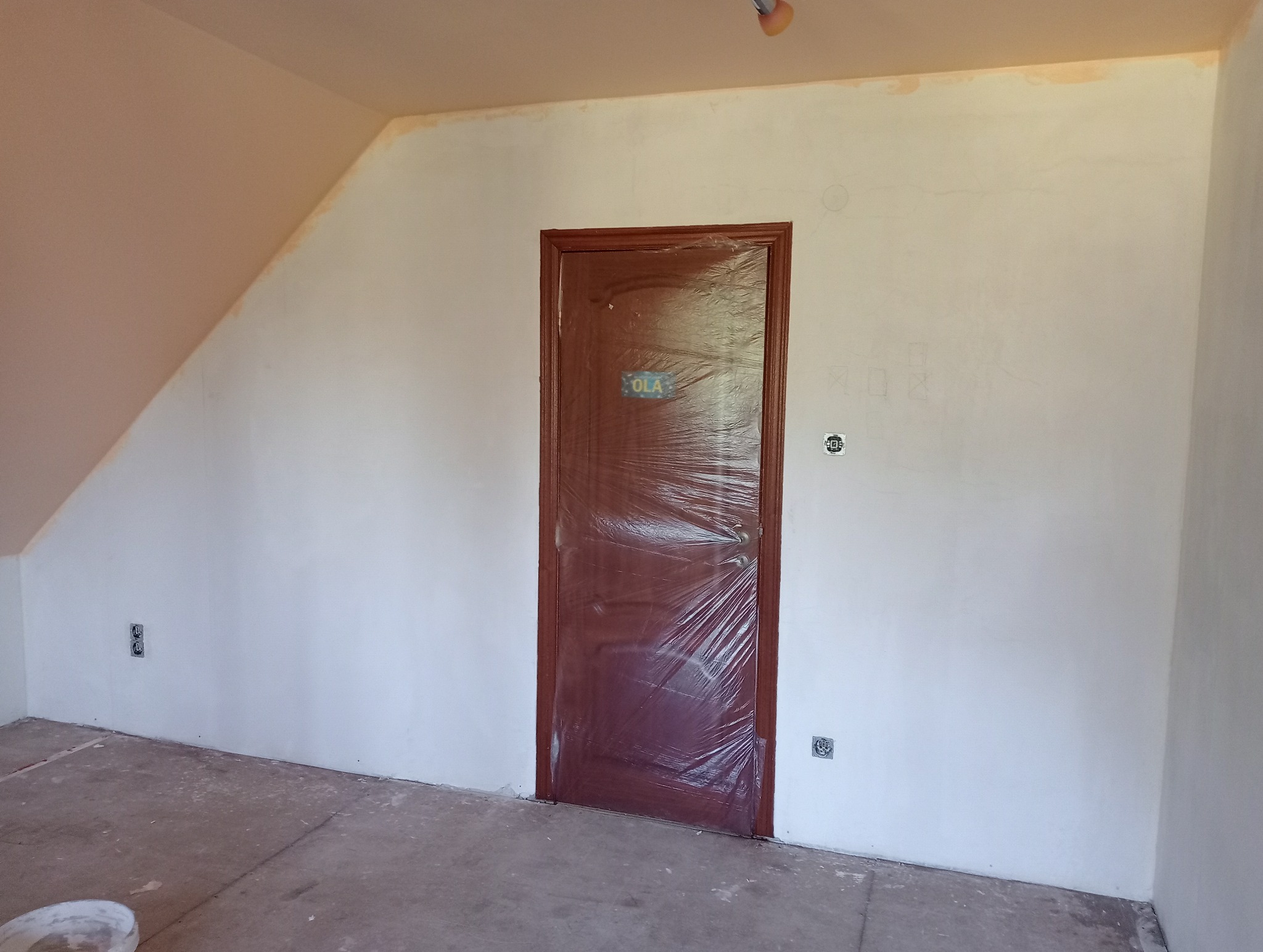 Pokój na poddaszu w trakcie prac przygotowawczych: ściany zagruntowane, zabezpieczone folią drzwi z tabliczką 'OLA', widoczne gniazdka elektryczne i ślady po ołówku na ścianach.