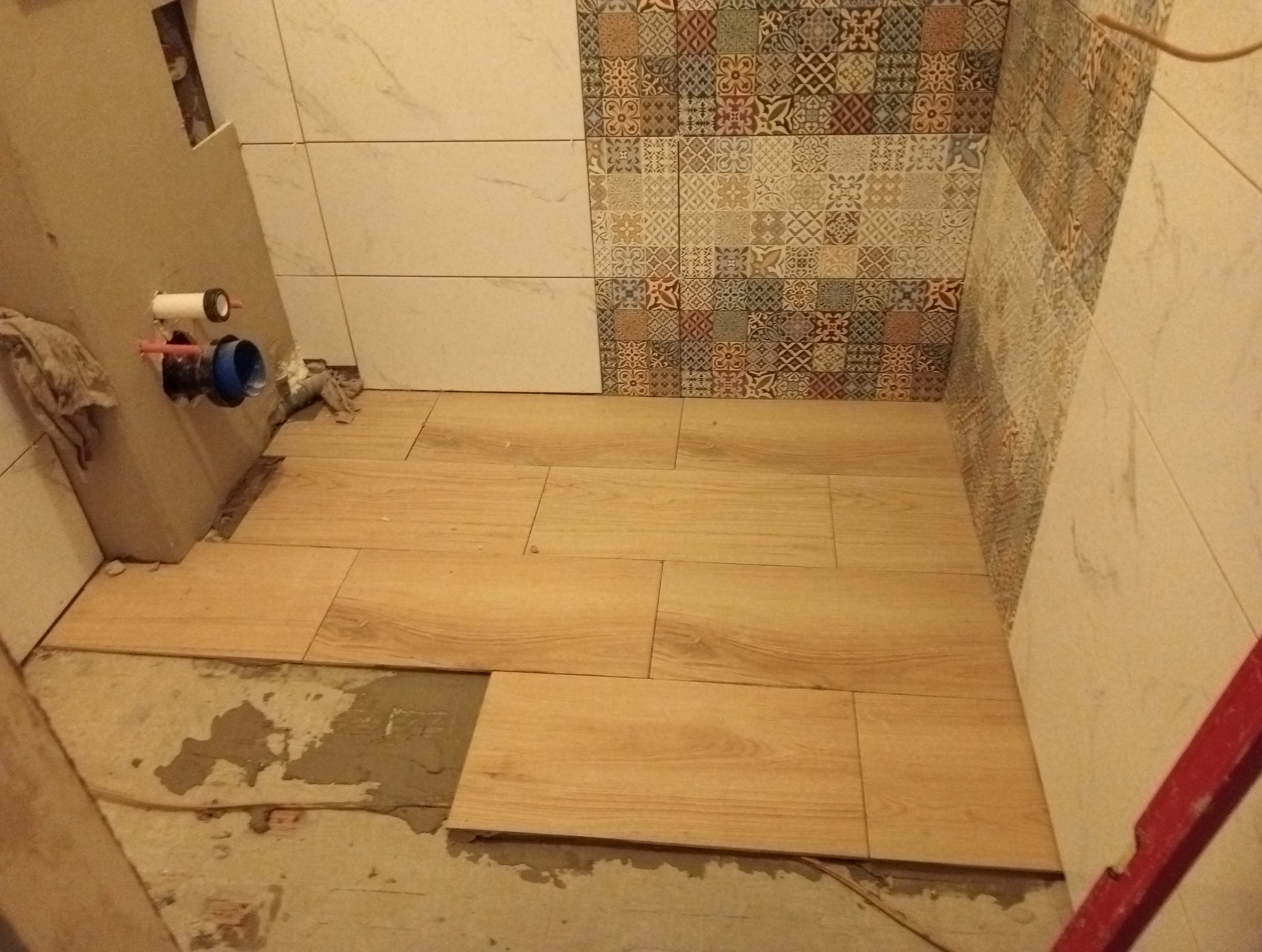 Układanie drewnopodobnych płytek podłogowych w łazience, widoczne fragmenty ściany z dekoracyjnymi i białymi płytkami oraz niezakończone prace przy instalacji sanitarnej.