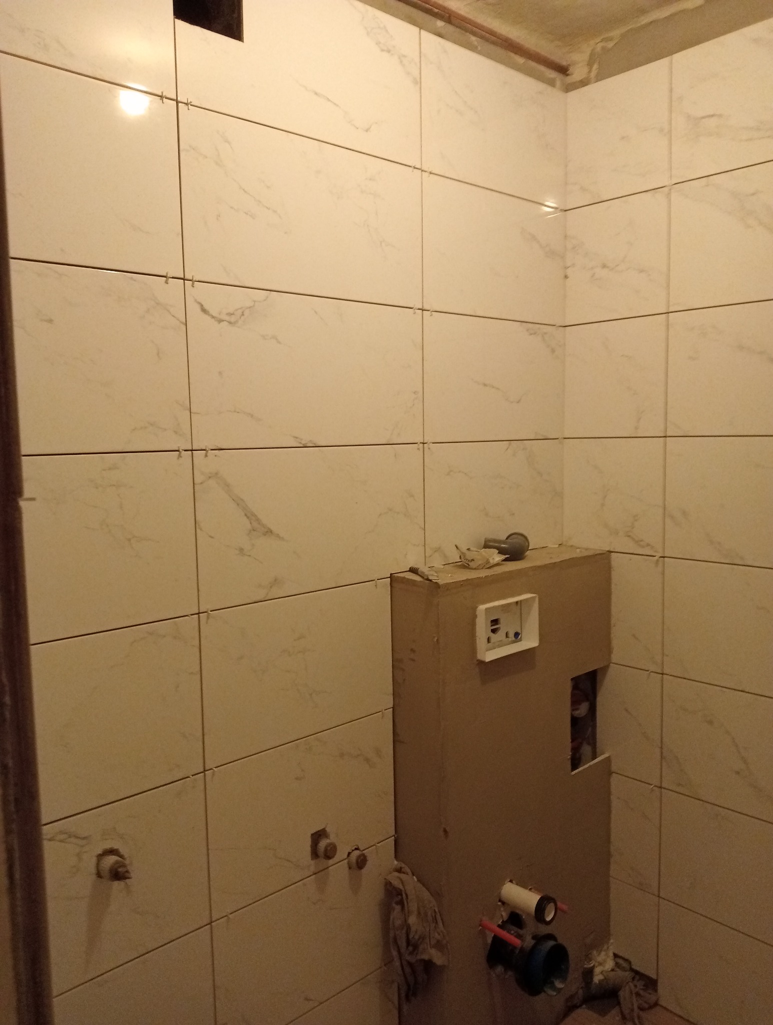 Ściana łazienki wyłożona jasnymi, marmurkowymi płytkami ceramicznymi, widoczne krzyżyki dystansowe w spoinach, zabudowa stelaża podtynkowego WC w surowym stanie, z widocznymi elementami instalacji.