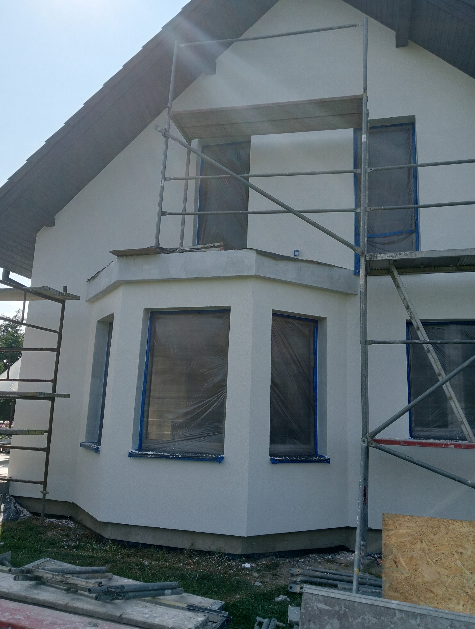 Dom jednorodzinny z oknami zabezpieczonymi folią i taśmą malarską, otoczony rusztowaniem podczas prac elewacyjnych w słoneczny dzień.
