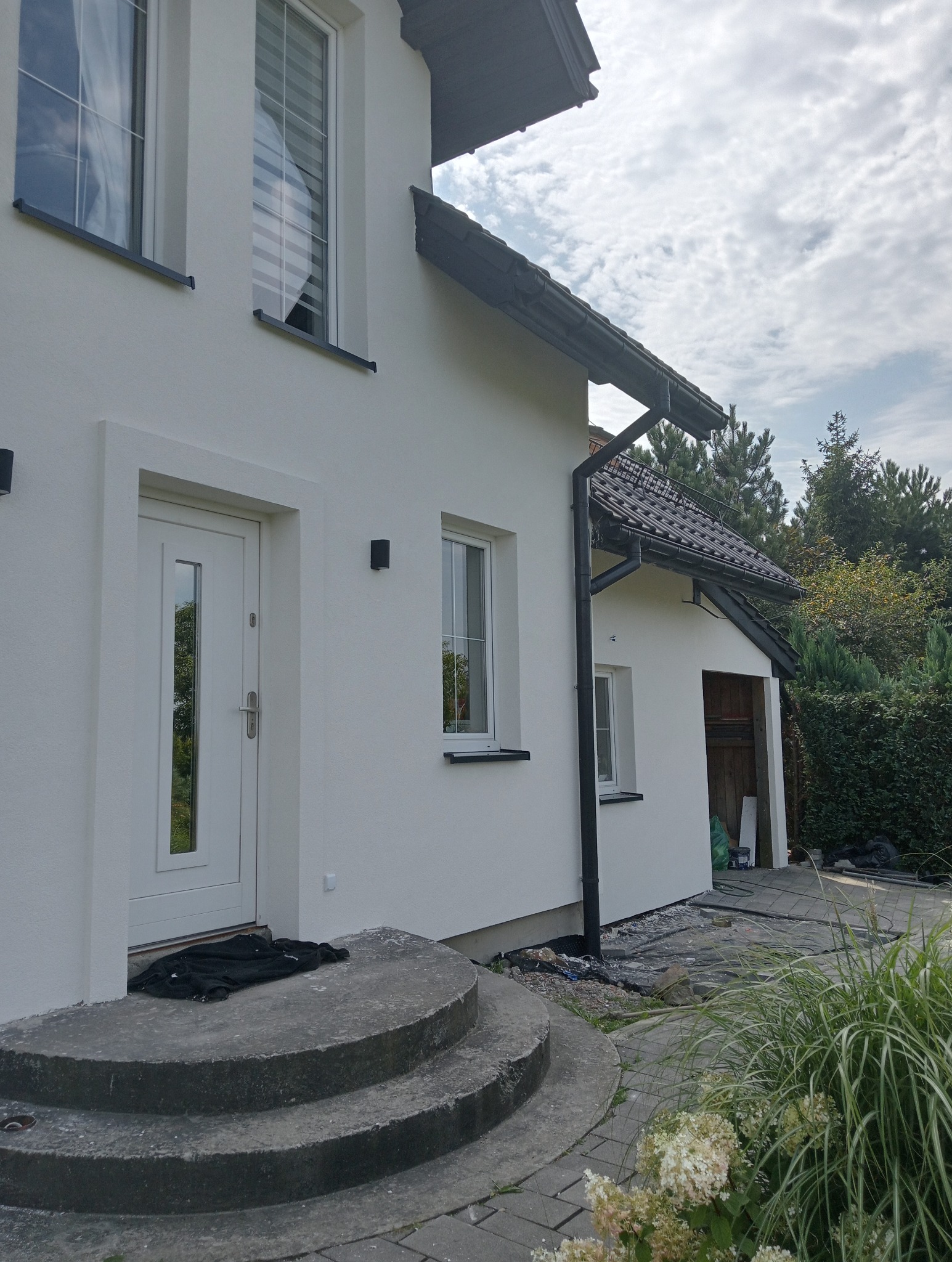 Biały dom z nową elewacją, widoczne okna z roletami, czarne rynny, schody wejściowe z czarnym materiałem i fragment ogrodu z roślinami.