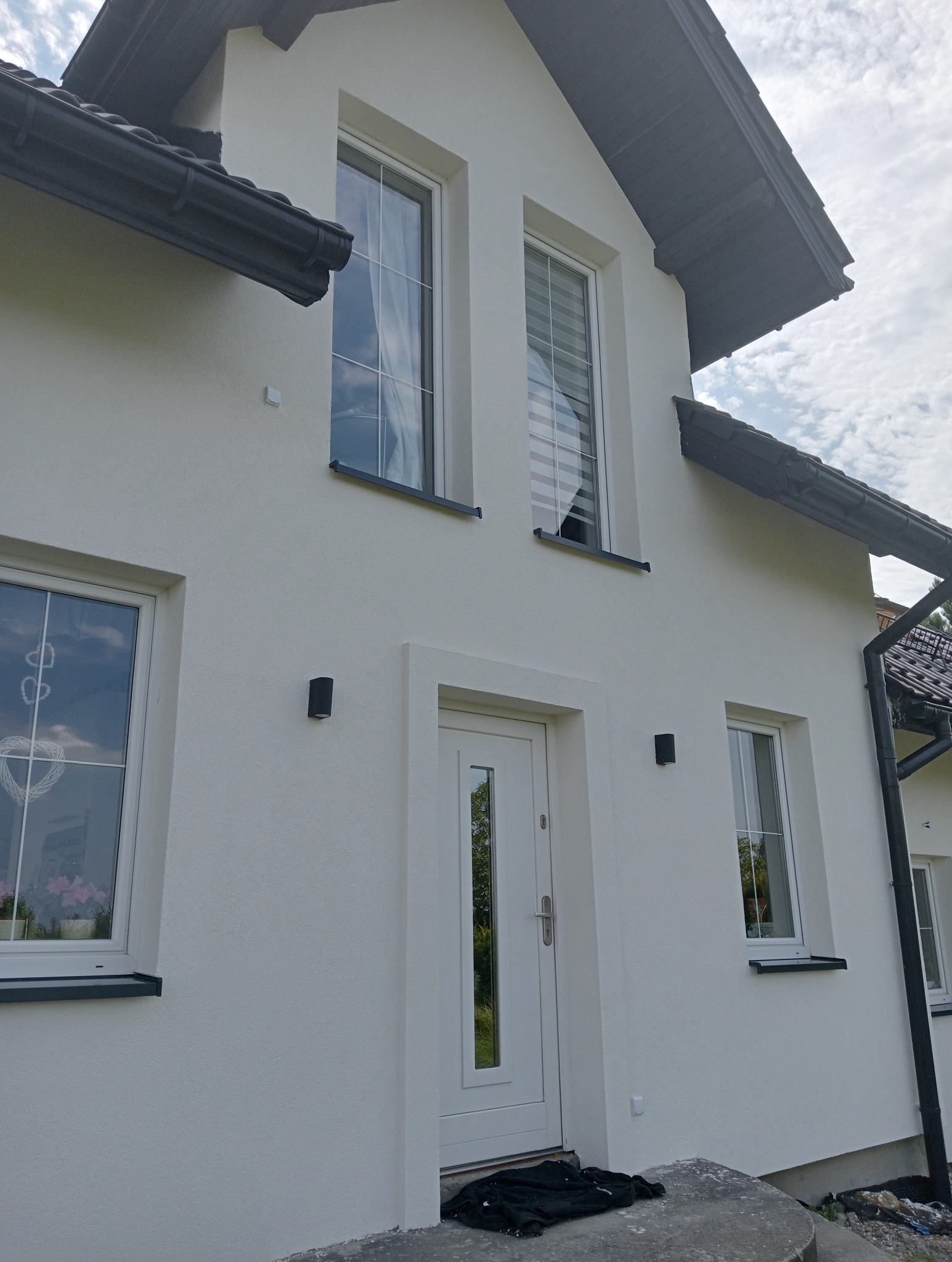 Biała fasada domu jednorodzinnego z widocznymi oknami z białymi ramami, drzwiami wejściowymi i czarnymi lampami elewacyjnymi, po zakończonym remoncie.