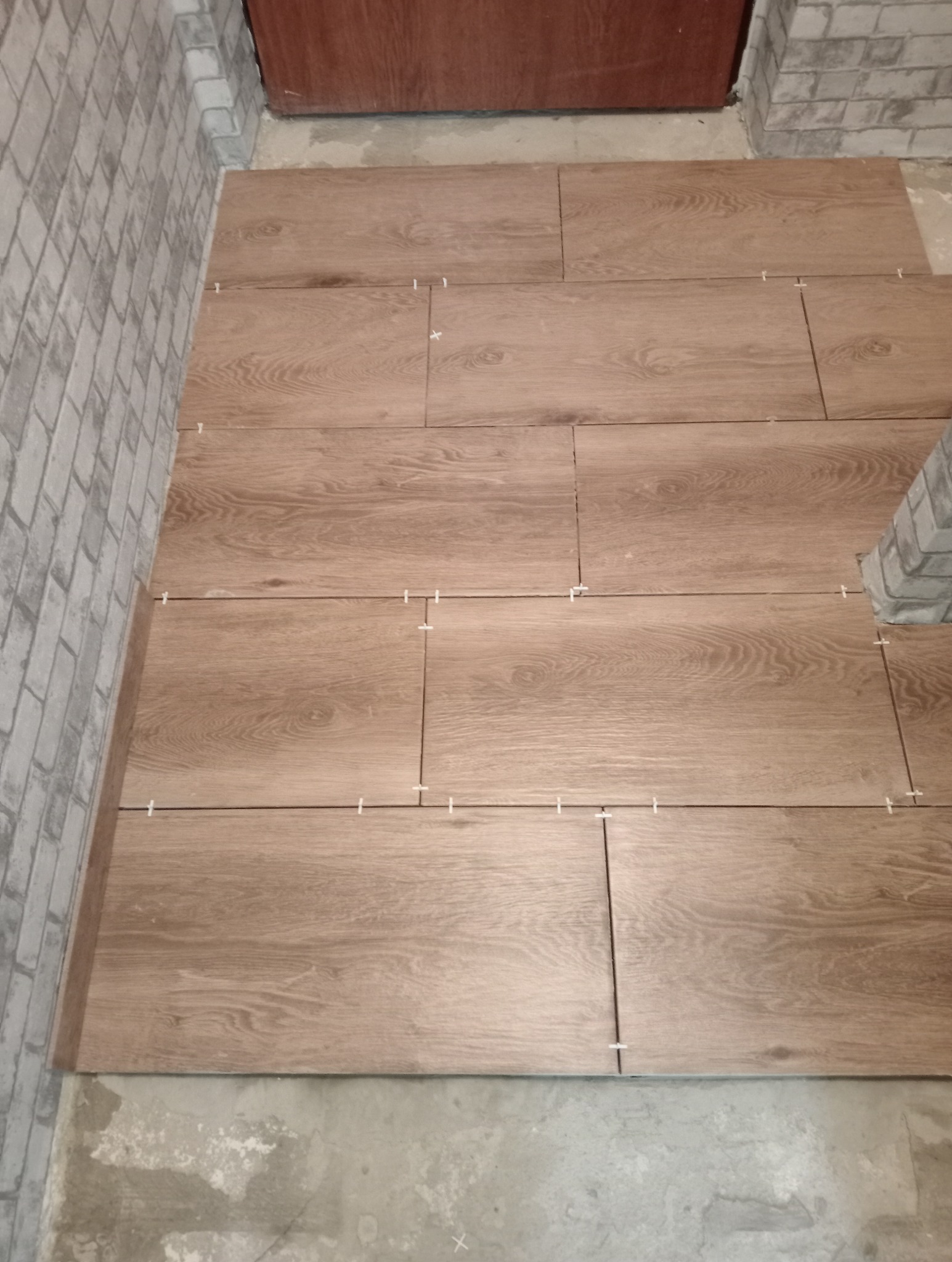 Układanie brązowych płytek podłogowych imitujących drewno z użyciem krzyżyków dystansowych, widoczna ściana z cegły i fragment drzwi.