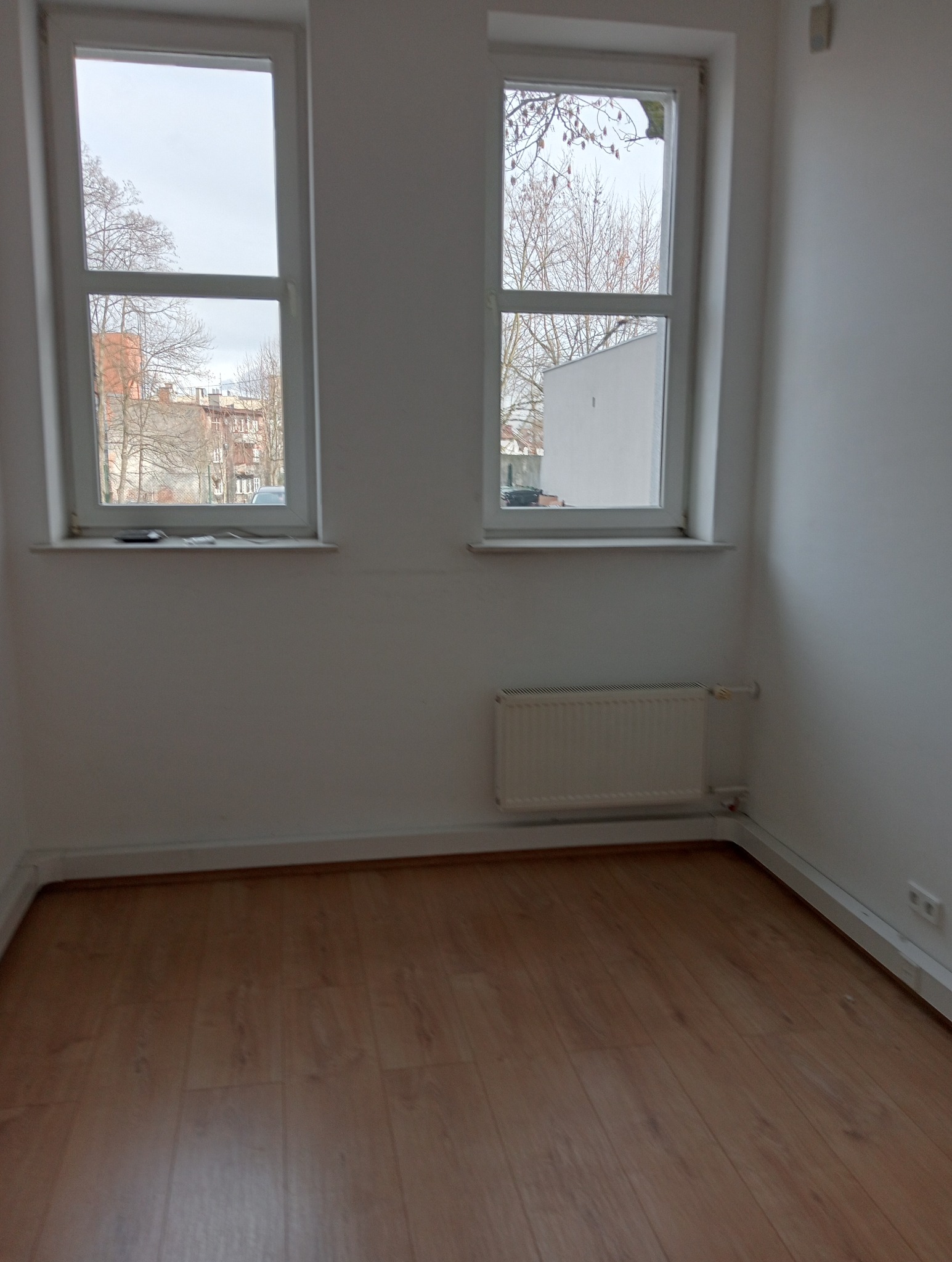Puste, białe pomieszczenie z dwoma oknami, drewnianą podłogą i białym grzejnikiem pod oknem
