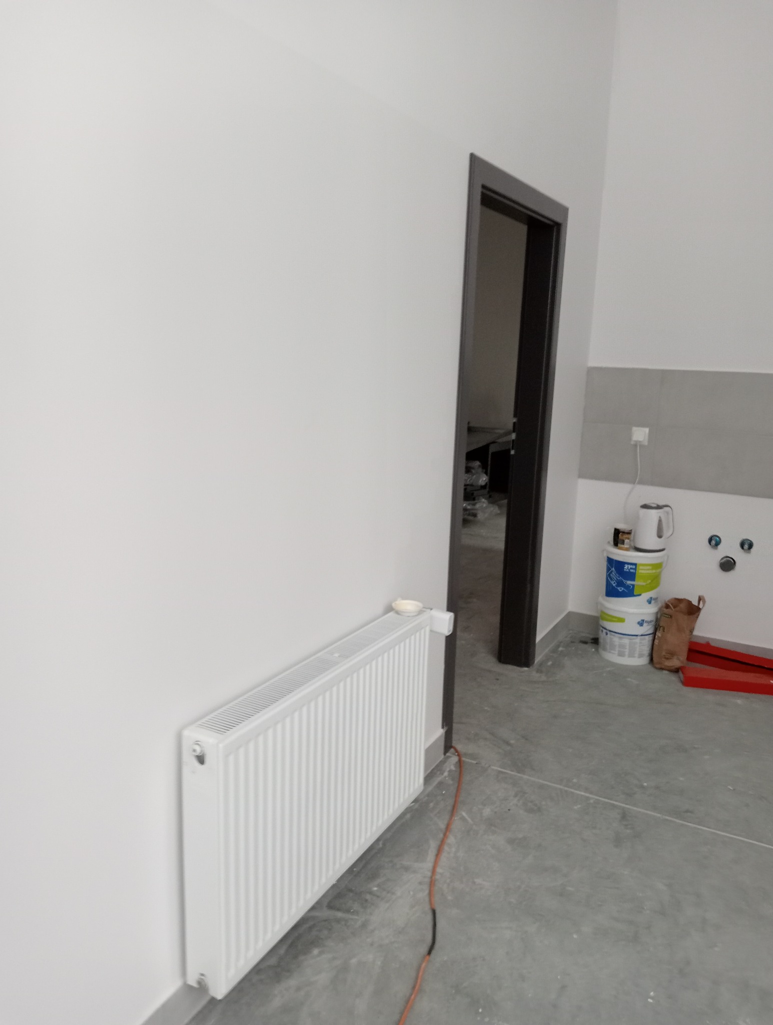 Świeżo pomalowane na biało ściany w pokoju, widoczny biały grzejnik, szara podłoga, czarna ościeżnica drzwi i wiaderka z farbą w tle.