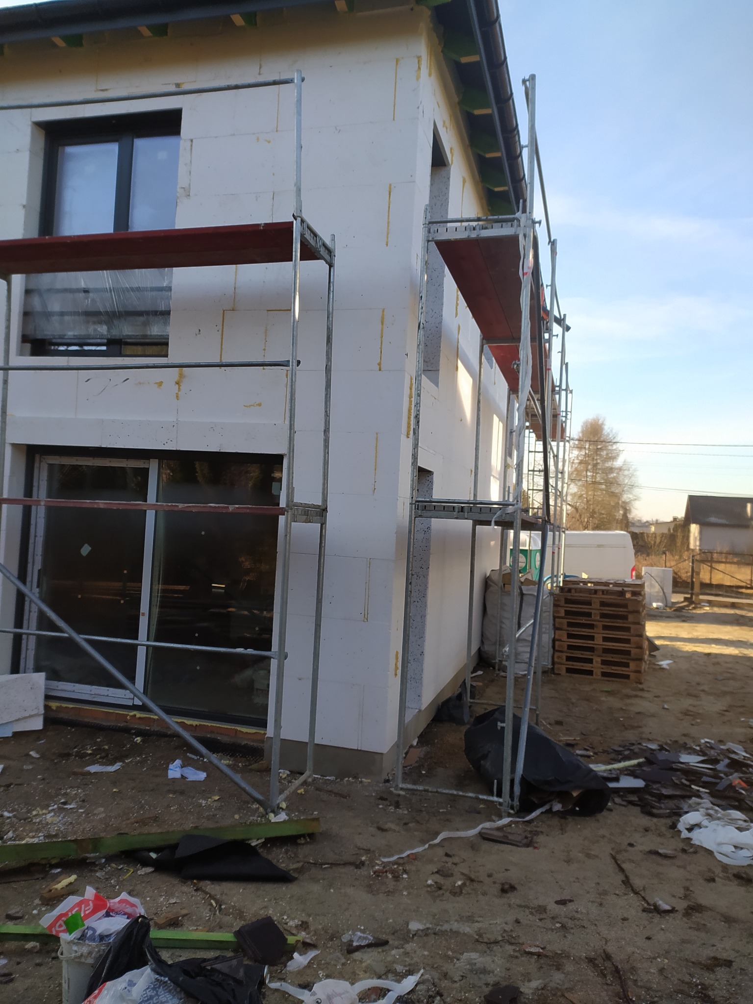 Dom w trakcie ocieplania, widoczne rusztowania, styropianowe płyty elewacyjne oraz palety i gruz na ziemi.