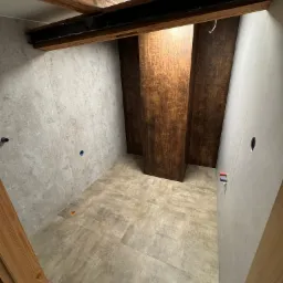 Wnętrze łazienki w trakcie remontu z szarymi płytkami na ścianach i podłodze, drewnianymi belkami stropowymi i pionowymi panelami w odcieniach brązu, widoczne otwory na instalacje.