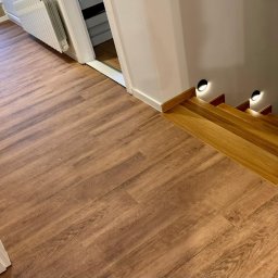 Silesia Boden - Ukończona podłoga z paneli winylowych o wzorze drewna, prowadząca do drewnianych schodów z oświetleniem punktowym i ceglanej ściany z gniazdkiem elektrycznym.