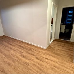 Silesia Boden - Jasna, pusta przestrzeń z nową podłogą imitującą drewno ułożoną w jodełkę, widok na okno i wejście do łazienki.