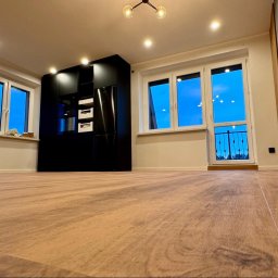 Silesia Boden - Jasne panele podłogowe w nowym mieszkaniu z widokiem na nocne niebo za oknem balkonowym i czarną zabudową kuchenną w tle.