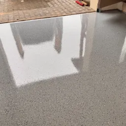 Błyszcząca, szara posadzka z drobnymi, kolorowymi wtrąceniami, odbijająca światło w garażu z otwartą bramą na zewnątrz.