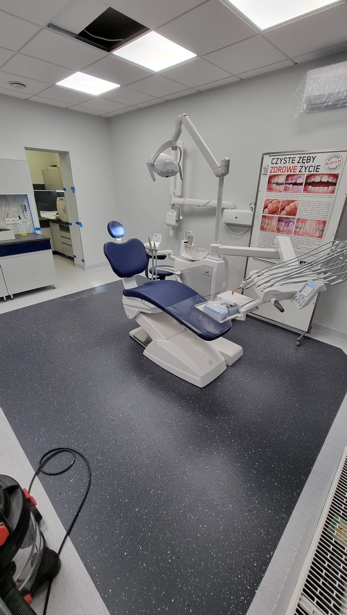Nowoczesny gabinet stomatologiczny w Izbicku z ciemną posadzką w drobne plamki, niebieskim fotelem dentystycznym i plakatem edukacyjnym o higienie jamy ustnej.