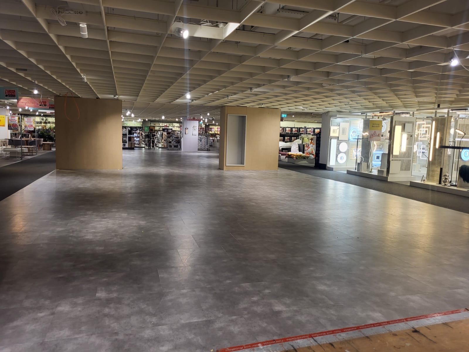 Szeroki kadr na szarą posadzkę w sklepie z widoczną kratownicą sufitową i regałami w tle.